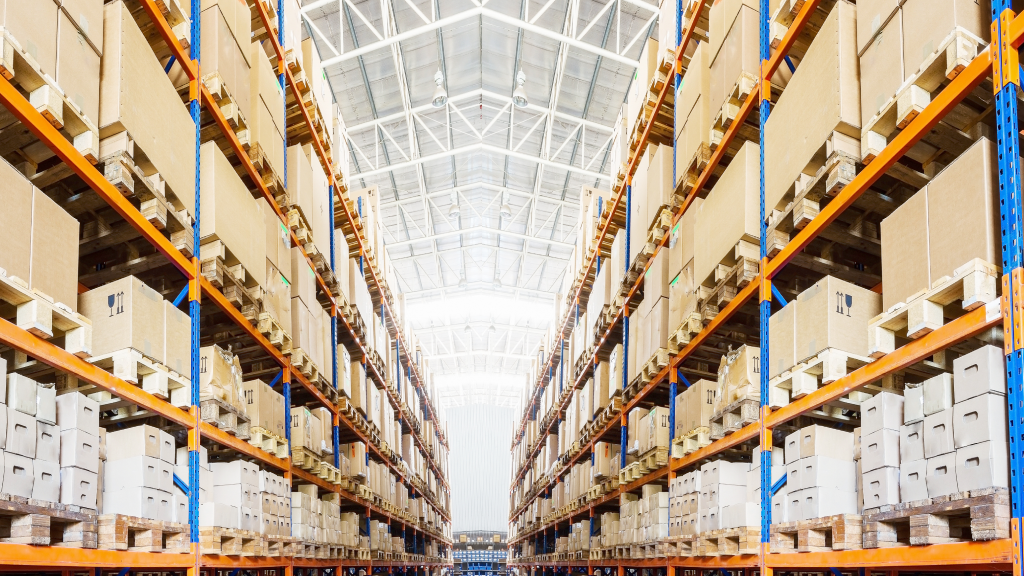 An image depicting a brightly lit warehouse aisle. The ceiling is high and the shelves a full with large boxes. 