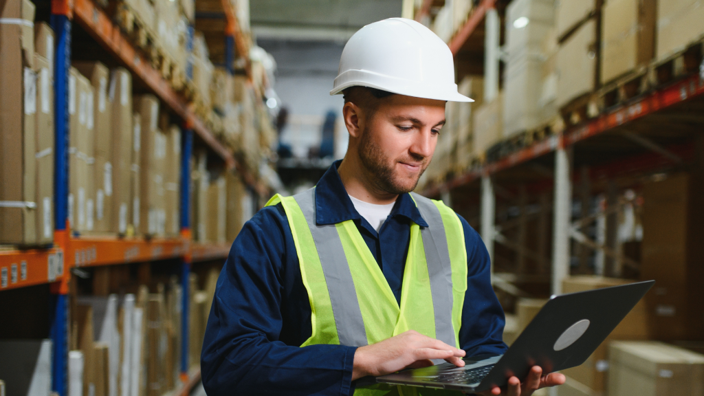 A warehouse worker stands in the middle of a a warehouse aisle working on their laptop.
