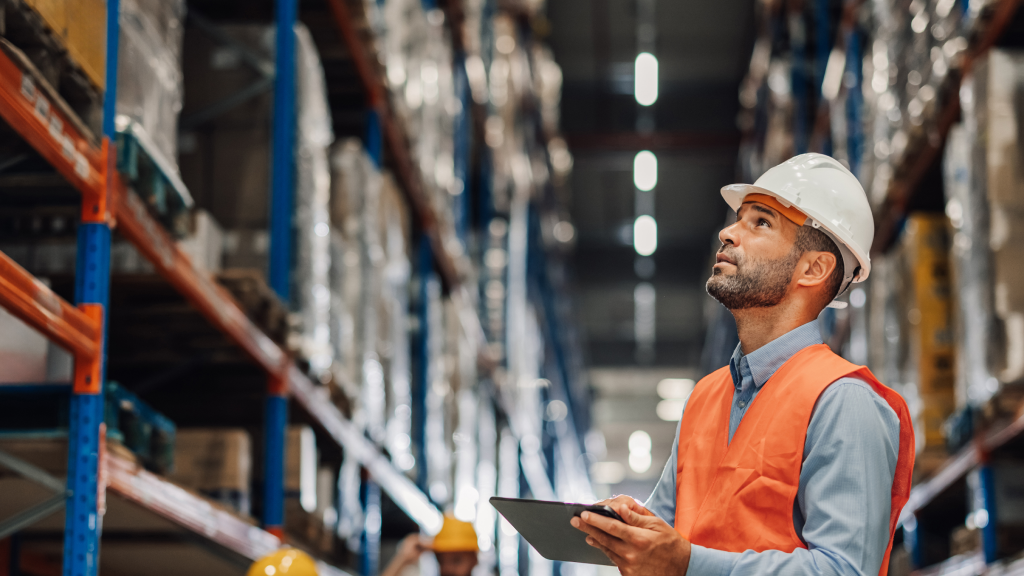 An image of a warehouse worker holding a tablet looking at the high racking above to find a specific item in the warehouse.