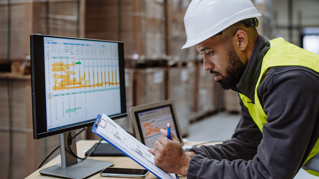 A warehouse worker cross-references his clipboard with what he can see on a monitor in front of him.