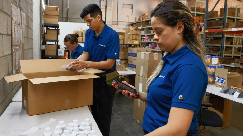 An image showing warehouse workers working in the warehouse. One packs orders while another checks an order on a mobile device.