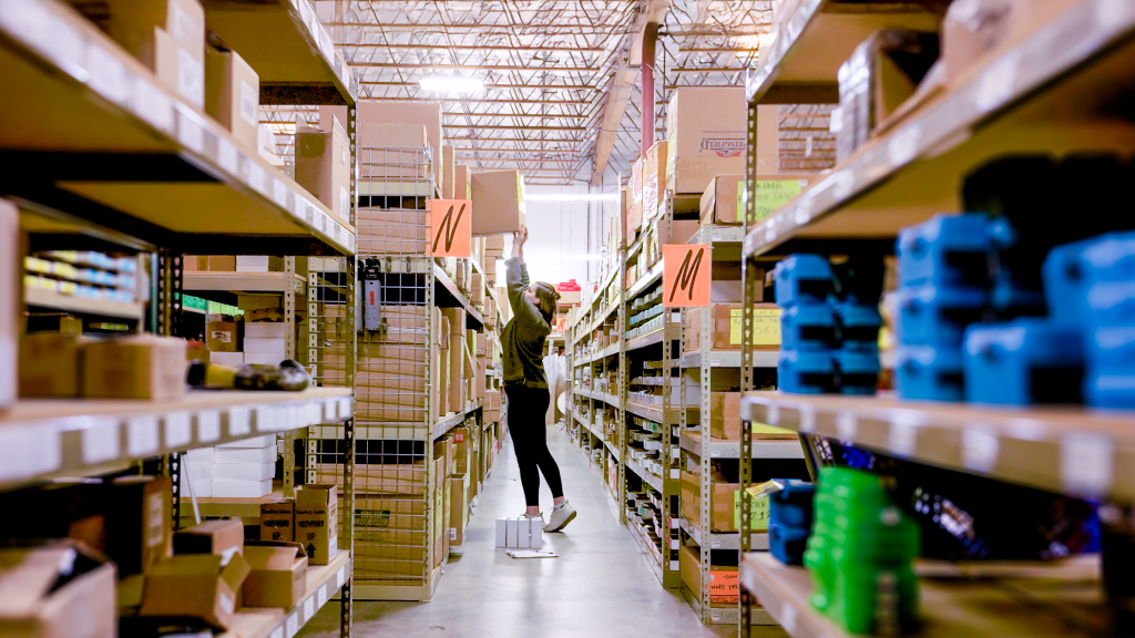 An image showing a warehouse worker lifting an order item from a shelf in the middle of an aisle.